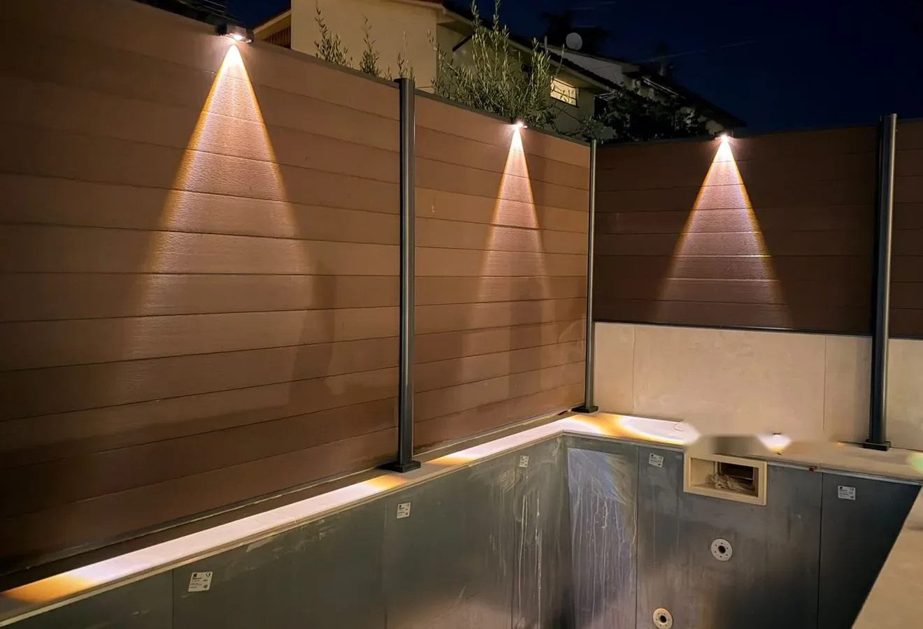 Piscina con bordo in gres chiaro e recinzione frangivista illuminata di sera